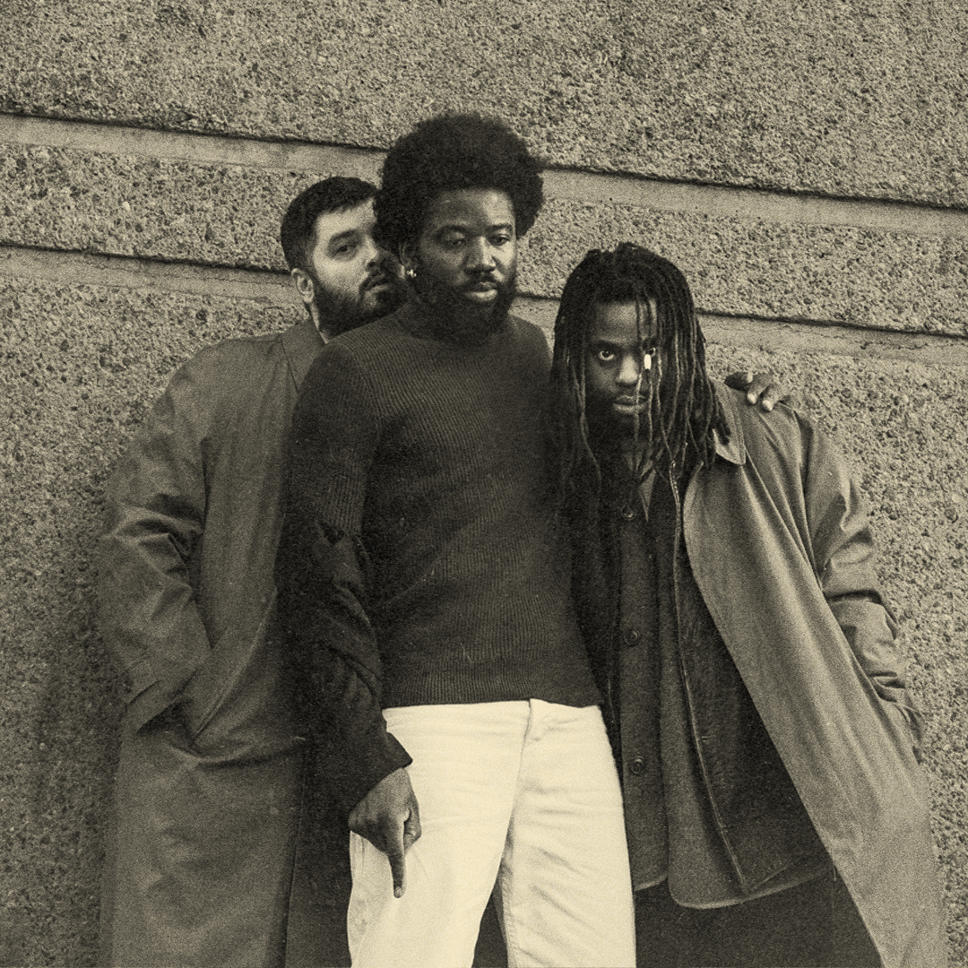 Black and white image of the three memebers from Young Fathers. The man on the left is wearing a long trench coat. The man in the middle has a black and white trousers. He's looking down and pointing his finger. The man on the right is looking into the camera, his hair hanging over his face. He's wearing a long jacket, with a shirt underneath. 
